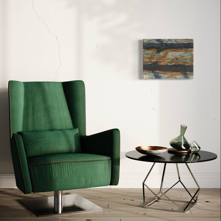 The artwork 'Elbows' by Emna Zghal features abstract horizontal bands of rustic blues and warm copper tones in a woodcut print on paper mounted on canvas, displayed above a modern emerald green armchair and round black side table, creating a striking contemporary interior composition. By Emna Zghal.