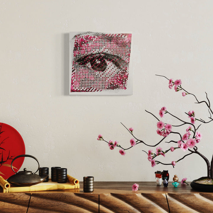 The artwork 'Pixelated' by Kiana Honarmand features a striking halftone-style eye rendered in black and pink dots against a white background. Hand-cut and woven digital prints create a mosaic-like effect, with delicate pink floral elements adorning the corners, measuring 16x16 inches. The composition merges traditional weaving techniques with contemporary digital art aesthetics.