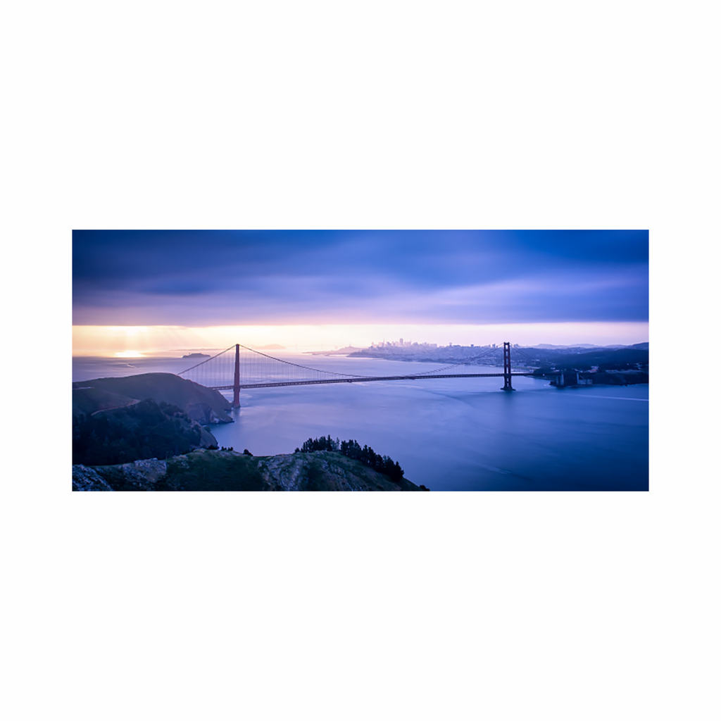 The artwork 'Full Bridge #2' by Dan Goldberger captures the Golden Gate Bridge in a dreamy panoramic view at twilight. The iconic suspension bridge spans across calm blue waters against a purple-hued sky, with San Francisco's skyline visible in the misty distance. Rolling hills and coastal vegetation frame the foreground, creating a serene coastal landscape bathed in soft evening light. Photograph by Dan Goldberger.