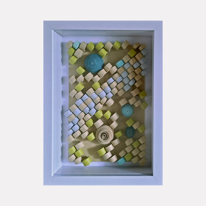 The artwork "Quilled" by Johanna Ryan features a framed 3D composition of small, rolled and folded paper pieces in shades of blue, green, cream, and beige, arranged in diagonal and spiral patterns that create a textured, geometric design with a delicate, handcrafted quilling style, highlighting intricate paper craft details.