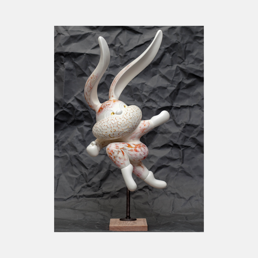 HOPE SPRINGS ETERNAL series by Yusuke Ochiai features a whimsical resin sculpture depicting a playful rabbit in mid-leap. The figure combines white resin with delicate orange-gold patterns and 22k gold leaf accents, set against a textured background. The sculpture's dynamic pose captures movement and joy, measuring 14x24x17 inches.