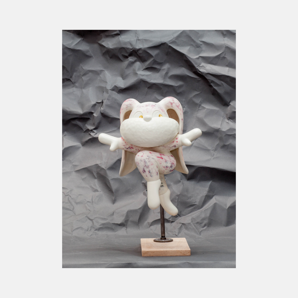 The artwork 'Leap of Faith' by Yusuke Ochiai features a whimsical white resin sculpture of a cheerful cat figurine balanced on a wooden base. The cat wears a delicate pink-patterned dress and spreads its arms wide in a joyful pose, enhanced with 22k gold leaf accents against a textured grey background. The sculpture embodies playful movement and artistic grace.