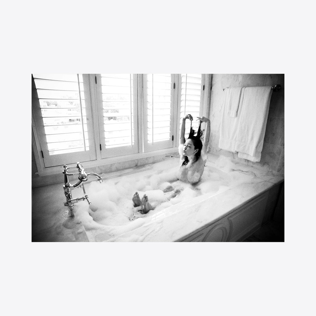 The artwork 'Rinka in the Morning Light' captures a tranquil bathroom scene in black and white photography. A vintage clawfoot bathtub filled with bubbling foam is set against a bright window with white shutters. The marble surroundings and classic faucet create an elegant, timeless atmosphere. A fine art archival pigment print by Mari Sarai.