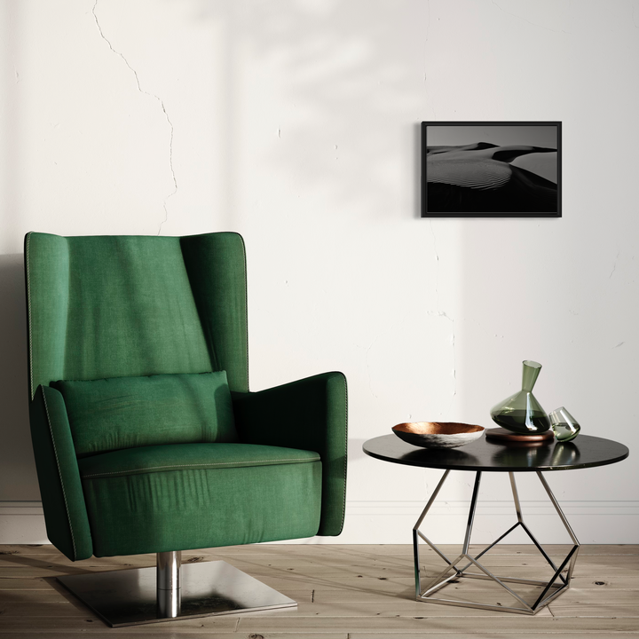 The artwork 'Pismo Beach Sand Dunes' by Neil Shapiro features a dramatic black and white photograph of undulating desert sand dunes. The monochromatic image captures the interplay of light and shadow across smooth, curved sand formations, creating a minimalist composition displayed in a black frame against a white wall. Interior setting shows modern decor with emerald green armchair.