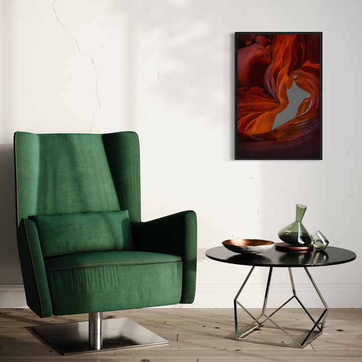 The artwork 'Slot Canyon In Color' by Neil Shapiro captures the dramatic swirling patterns of a sandstone canyon in vibrant orange and red hues. The metal print showcases smooth, flowing curves resembling liquid fire against a deep burgundy background, displayed in a modern room setting with a green velvet chair and contemporary round table.