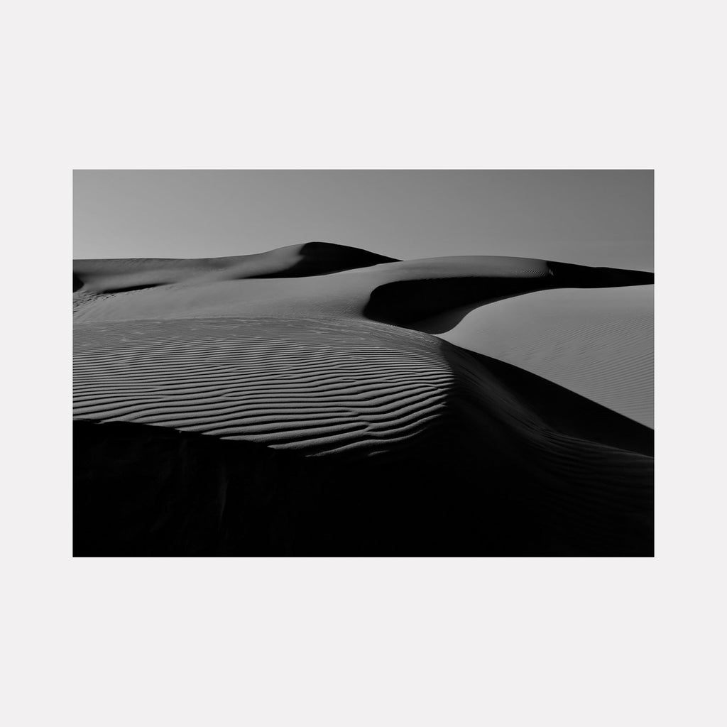 The artwork 'Pismo Beach Sand Dunes' by Neil Shapiro captures dramatic black and white desert landscape featuring undulating sand dunes with distinct ripple patterns. Strong contrast between light and shadow creates mesmerizing geometric patterns across the rolling dunes, showcasing natural textures in monochromatic photography printed on Moab Entrada paper.