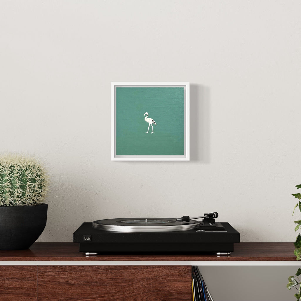 The artwork 'Profit Margins, Flamingo' by Jacki Davis features a minimalist white flamingo silhouette centered on a seafoam green background, framed in white. Created using US currency and acrylic on wood panel, the 8x8 inch piece hangs above a vintage turntable, offering a striking contrast against a neutral wall. By Jacki Davis.