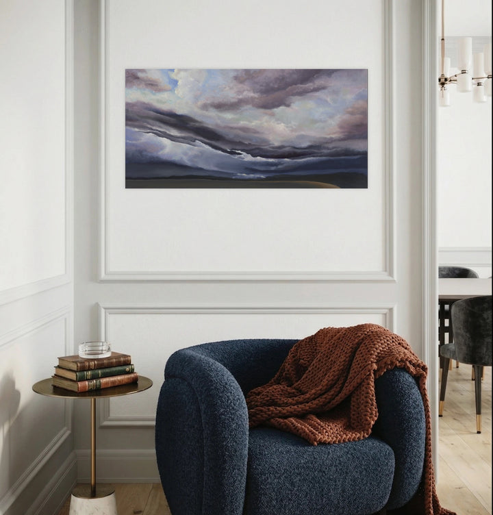Muted Purple Sky by Nadia Parsons reveals a sweeping landscape beneath textured clouds blending soft purples and muted blues, framed in a serene modern interior featuring a deep blue armchair and warm brown knit throw, evoking calm and contemplative artistic ambiance.