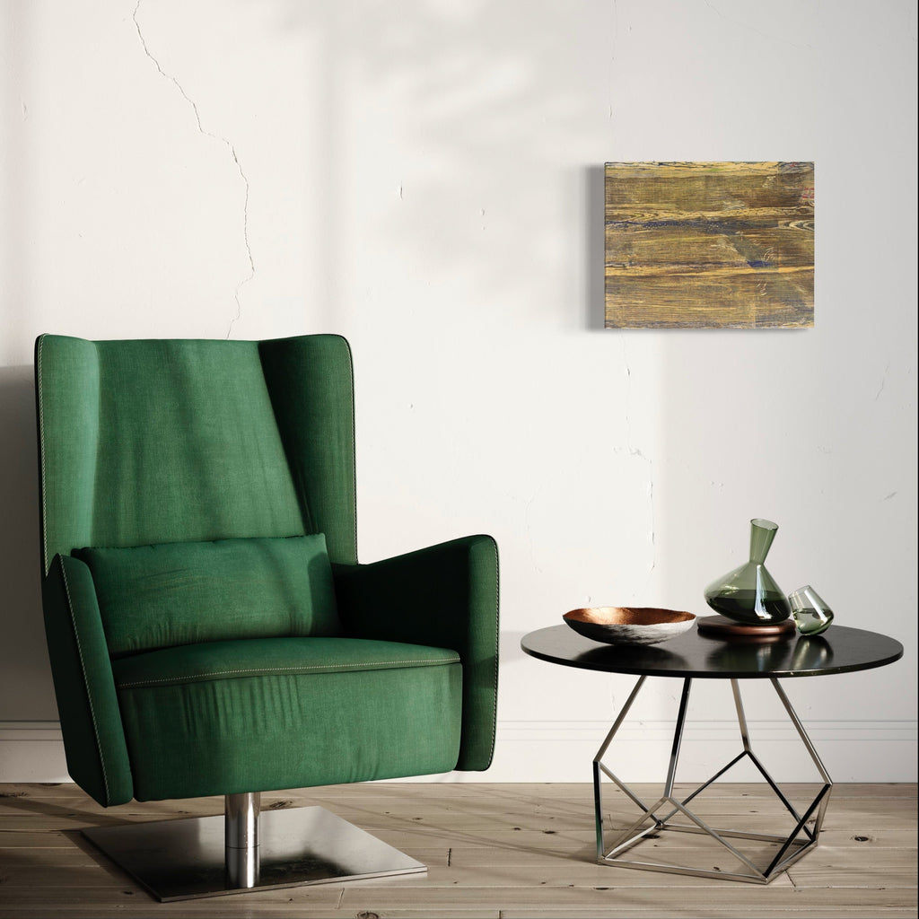 The artwork 'Floating Core' by Emna Zghal features abstract woodcut patterns on paper mounted on canvas, displaying textural layers in earthy golden-brown tones. Natural wood grain textures create a harmonious composition with horizontal striations, suggesting organic movement and depth, mounted on a clean white wall above a modern green velvet armchair.