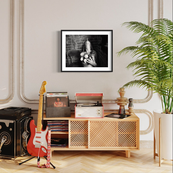 The artwork 'Rose & Cigarette' by Mari Sarai features a striking black and white archival pigment print in a sleek black frame. The 20x24 inch composition displays powerful contrast and intimate documentary-style photography. The print is shown in a modern interior setting above a vintage-style wooden credenza, complemented by surrounding musical elements and greenery.