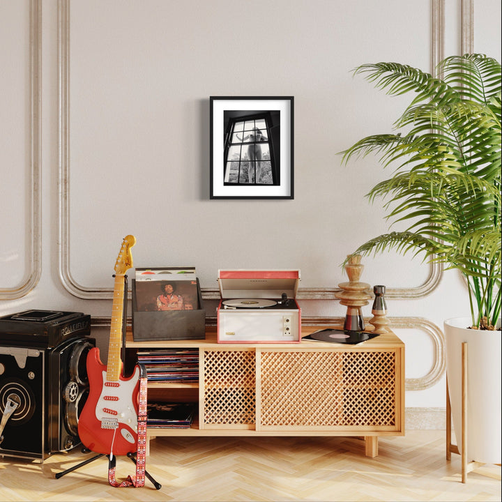 The artwork 'Girl in the Wonderland' by Mari Sarai features a black and white framed photograph displayed in an artistic living space, mounted on a beige wall. Set against a retro-modern interior with herringbone flooring, vintage record player, wooden lattice console, red electric guitar, and lush palm plant. Archival pigment print, 16.5 x 12 inches, by Mari Sarai.