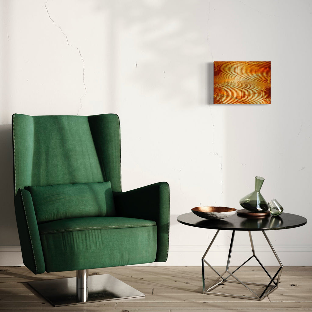 The artwork 'Land of Sunshine 01' by John Cox features vibrant concentric circles in warm orange and gold tones, displayed on a canvas above a modern green armchair. The abstract piece showcases dynamic wave-like patterns creating a sense of movement and energy through textured acrylic brushstrokes on a 12x16 inch canvas. By John Cox.