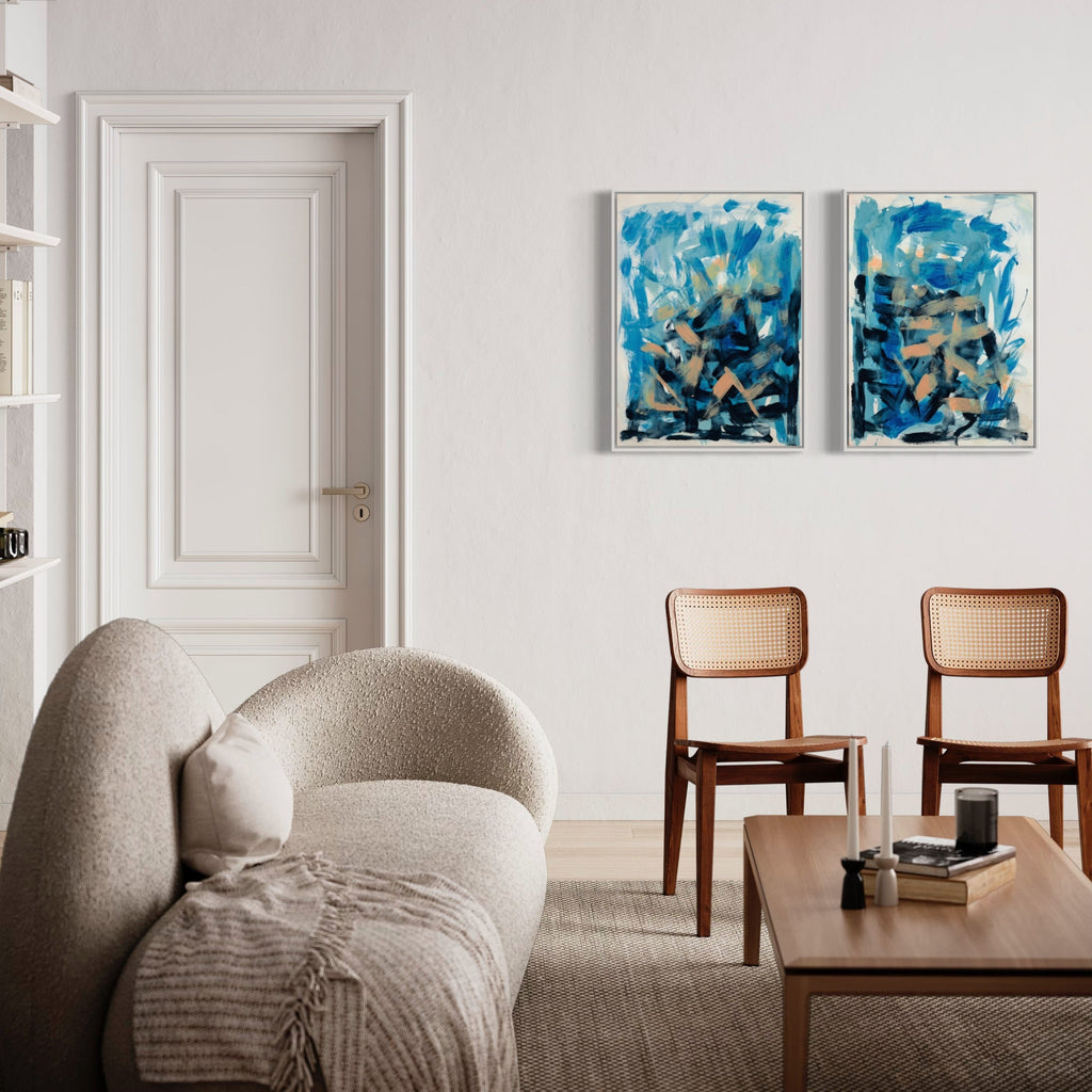 The artwork 'Chimera' by Molly Herman features a diptych of abstract paintings with dynamic brushstrokes in vibrant turquoise and deep navy blues, contrasted with earthy neutrals. Each panel displays expressive gestural marks creating a sense of movement and depth on BFK Rives paper, framed in complementary white frames. Artwork by Molly Herman.