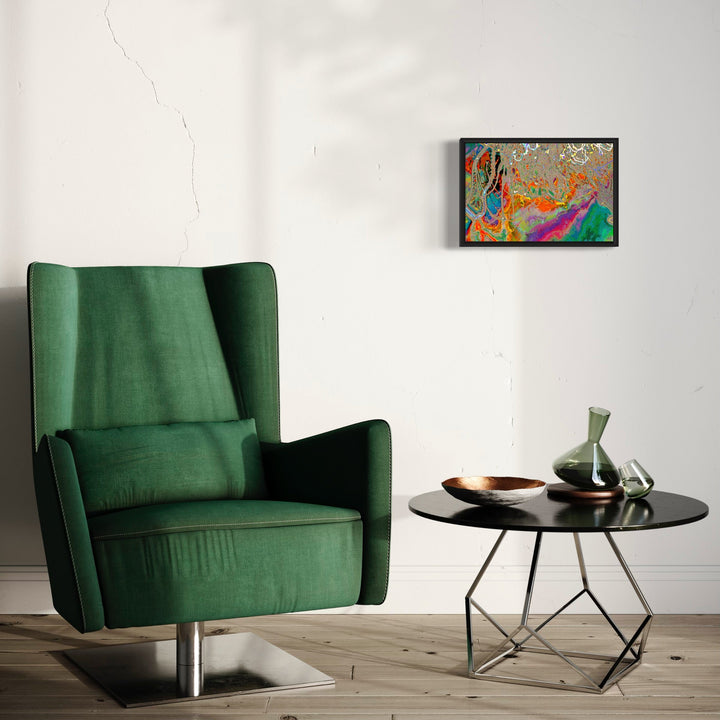 The artwork 'Particle Physics 18' by Chunbum Park features vibrant abstract patterns in orange, blue, and magenta hues against a textured background, displayed in a sleek black frame. The modern giclee print showcases dynamic swirling formations reminiscent of subatomic particle movements, creating a striking contrast against the room's emerald green armchair and minimalist decor.