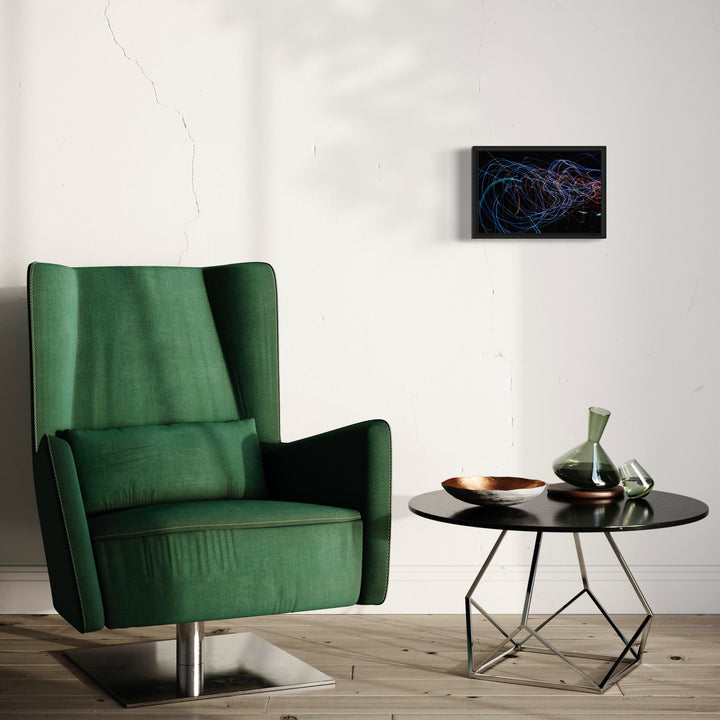 The artwork 'Particle Physics 90' by Chunbum Park features dynamic swirling lines in vibrant blue and red, creating an abstract visualization of particle movement against a black background. The small-format giclee print is displayed on a white wall above a modern interior setting with an emerald green armchair and round black side table with decorative vases. By Chunbum Park.