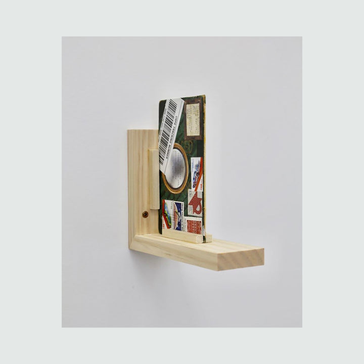 The artwork 'Bus to Chinatown' by Mitsuko Brooks features a mixed-media collage mounted on a light wooden shelf against a gray background. The piece incorporates vintage postage stamps, book cover elements, and typewritten text arranged in a vertical composition, with visible textures from rice paper and postal markings. - Mitsuko Brooks