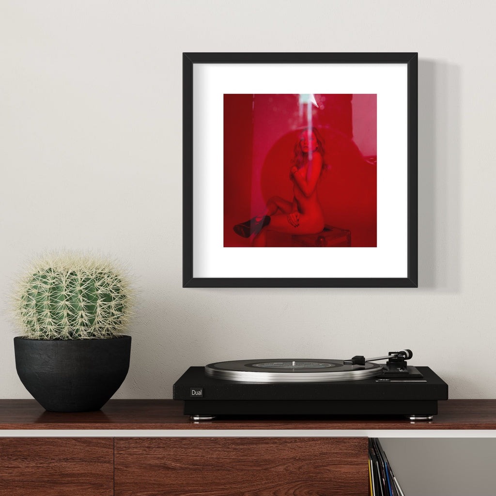 The artwork 'Clara in the Red Light' by Mari Sarai features a striking composition bathed in deep red lighting. A silhouetted figure appears contemplative against the vibrant crimson background, creating a dramatic and atmospheric effect. The archival pigment print is presented in a sleek black frame with white matting, creating a stark contrast with the intense red tones. 11x14 inches.