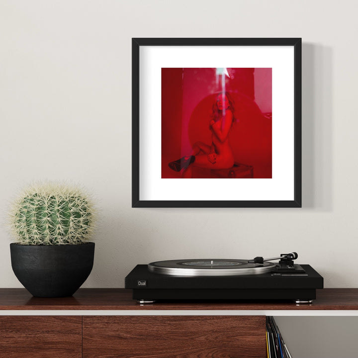 The artwork 'Clara in the Red Light' by Mari Sarai features a striking composition bathed in deep red lighting. A silhouetted figure appears contemplative against the vibrant crimson background, creating a dramatic and atmospheric effect. The archival pigment print is presented in a sleek black frame with white matting, creating a stark contrast with the intense red tones. 11x14 inches.