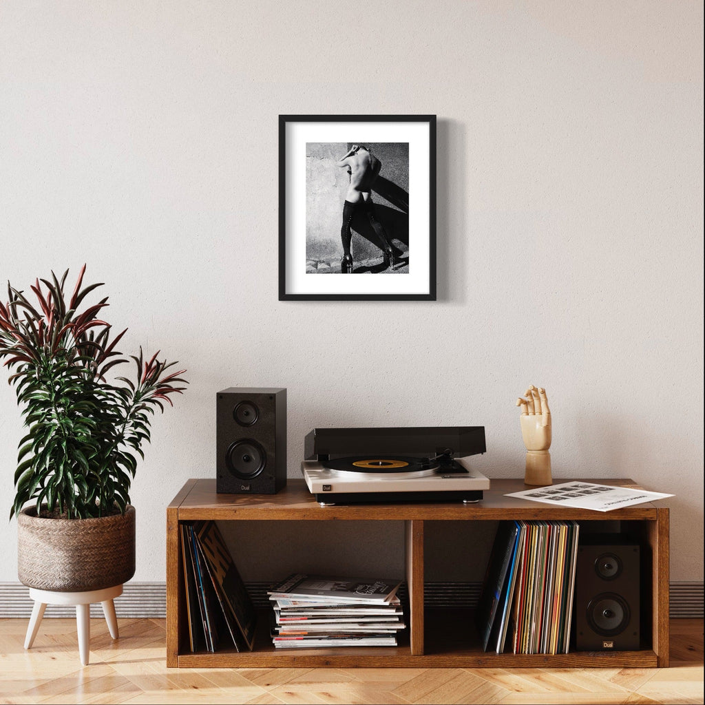 The artwork 'Cock Heel' by Mari Sarai features a striking black and white archival pigment print in a sleek black frame. The monochromatic composition shows a dramatic silhouette against a textured background, displayed above a vintage-style record console. The 11x14 inch print creates a bold artistic statement in a modern interior setting.