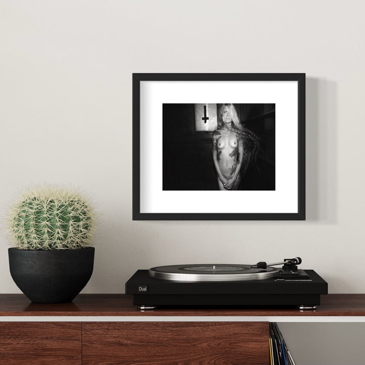 The artwork 'Fallen Metal Angel' by Mari Sarai features a black and white monochromatic composition in a stark, dramatic style. The ethereal form appears suspended against a dark background, with contrasting light and shadow elements. The archival pigment print is displayed in a classic black frame, emphasizing its contemporary artistic nature. By Mari Sarai.