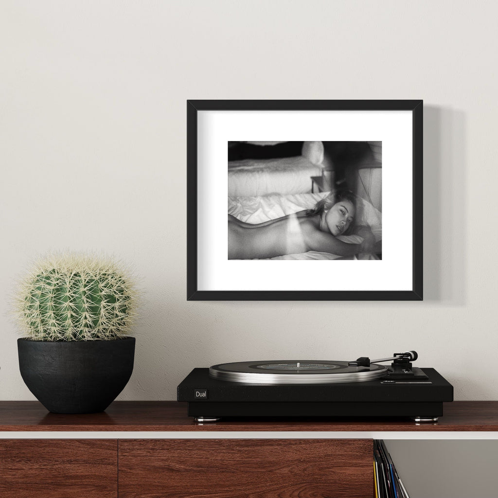 The artwork 'Hikari in Arizona Breeze' by Mari Sarai features a black and white archival pigment print displayed in an elegant black frame. The composition shows soft, ethereal tones with dramatic light and shadow interplay, creating a dreamlike atmosphere. The 11x14 inch print is matted in white, complementing the minimalist presentation by Mari Sarai.