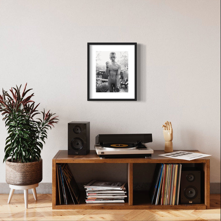 The artwork 'Let Me In!' features a black and white archival pigment print in a sleek black frame, displayed above a mid-century modern vinyl setup. The 11x14 inch print contrasts against a light neutral wall, creating a striking gallery-style presentation amid a vinyl collector's home setting with speakers and indoor plants. By Mari Sarai.