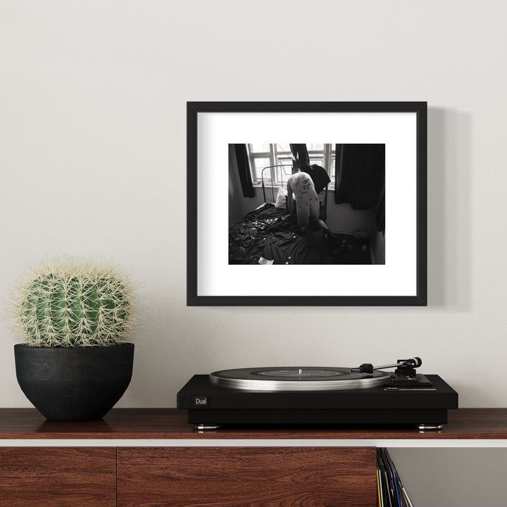 The artwork 'Money Bed' by Mari Sarai features a black and white archival pigment print in a sleek black frame, showing an interior scene with scattered objects near a window. The composition creates a moody atmospheric quality through dramatic lighting and shadows. Displayed alongside a potted cactus and turntable, measuring 11x14 inches. By Mari Sarai.
