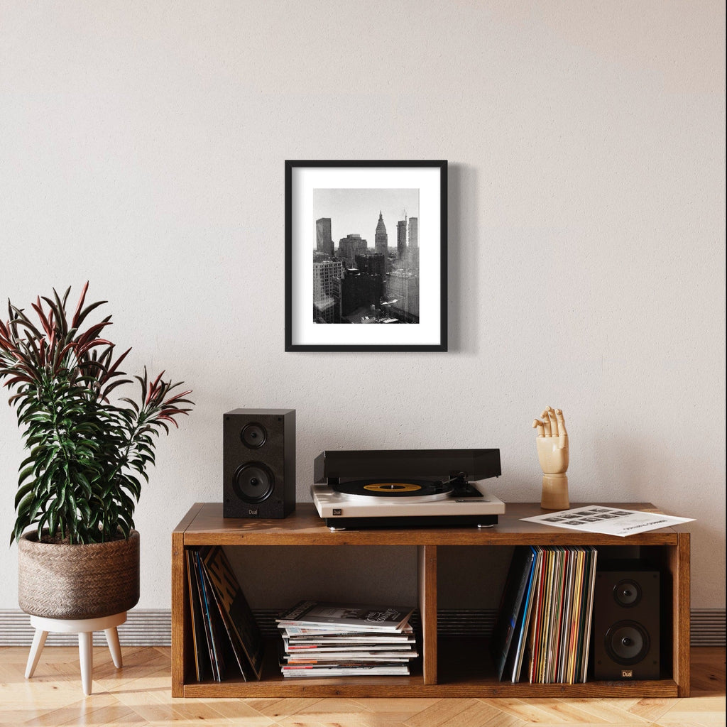 The artwork 'NY Melancholy' by Mari Sarai captures a moody black and white cityscape of New York's iconic skyline. The archival pigment print showcases towering skyscrapers silhouetted against a misty sky, including the distinctive Empire State Building. The composition presents a dramatic contrast between dark architectural forms and light atmospheric elements, framed elegantly in black. Photographed with artistic sensitivity by Mari Sarai.