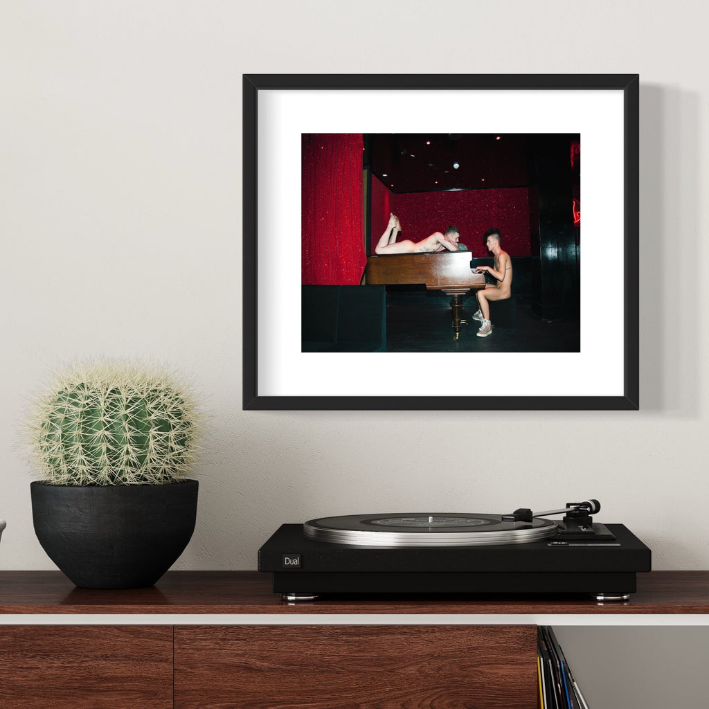 The artwork 'Piano Piano' features a grand piano against a deep red backdrop in a dimly lit space. The musical instrument, rendered in archival pigment print, creates a dramatic composition with strong contrast between dark and illuminated areas. The 11x14 inch framed piece showcases the elegant curves of the piano in a theatrical setting by Mari Sarai.