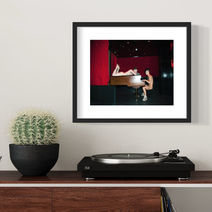 The artwork 'Piano Piano' features a grand piano against a deep red backdrop in a dimly lit space. The musical instrument, rendered in archival pigment print, creates a dramatic composition with strong contrast between dark and illuminated areas. The 11x14 inch framed piece showcases the elegant curves of the piano in a theatrical setting by Mari Sarai.