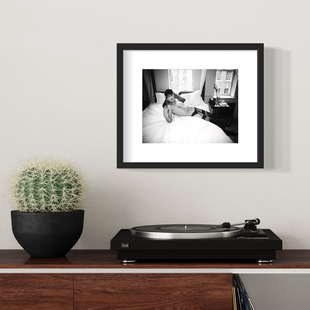 The artwork 'Pistol & Bed' by Mari Sarai features a monochromatic composition showing a reclining figure on white bedding, captured in black and white. The archival pigment print is presented in a classic black frame, displaying dramatic contrast and intimate documentary-style photography.