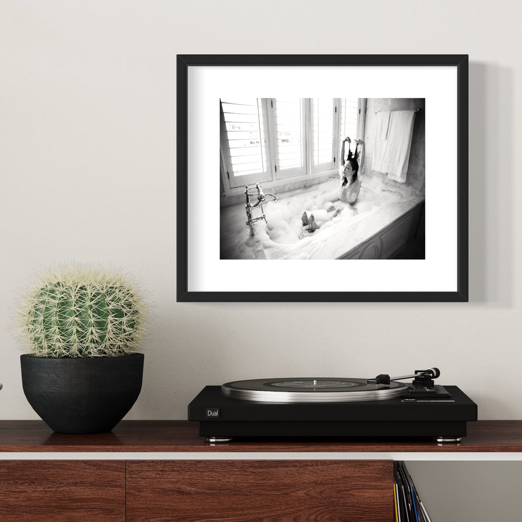 The artwork 'Rinka in the Morning Light' captures a tranquil bathroom scene in black and white photography. A vintage clawfoot bathtub filled with bubbling foam is set against a bright window with white shutters. The marble surroundings and classic faucet create an elegant, timeless atmosphere. A fine art archival pigment print by Mari Sarai.