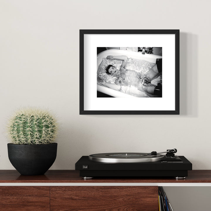 The artwork 'Simulated Experience' features a black and white archival pigment print in a sleek black frame. The composition shows a bathtub scene with splashing water creating dynamic patterns and reflective textures. The high contrast monochromatic style emphasizes the interplay of light and shadow across the water's surface. 11x14 inches print by Mari Sarai.