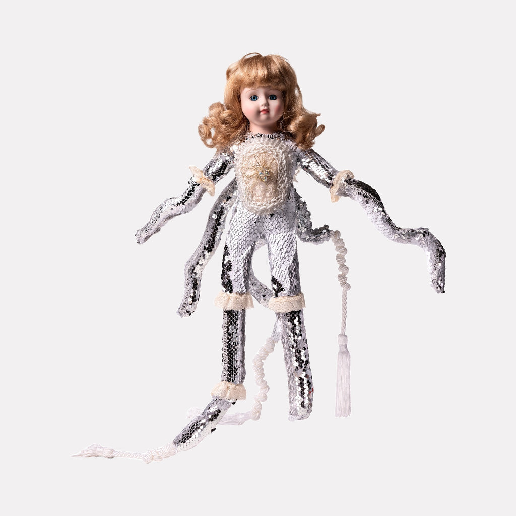 A surreal doll figure with porcelain face and curly hair, dressed in a textured costume with shimmering sequins and soft fabric details, featuring multiple elongated limbs. This unique creation is part of Lisyanet Rodriguez’s "A Worm, a Bird, an Insect, a Human Being…" art collection for Curina.