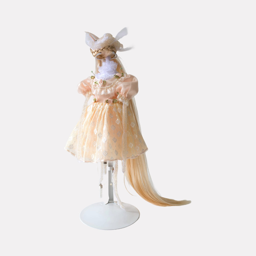 Mellow Fever by Lisyanet Rodriguez is a whimsical art piece featuring a delicate figure dressed in a soft peach dress adorned with floral accents, complemented by a long, flowing tail and intricate headpiece, blending imaginative craftsmanship with elegant detail for the Curina collection.
