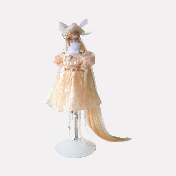Mellow Fever by Lisyanet Rodriguez is a whimsical art piece featuring a delicate figure dressed in a soft peach dress adorned with floral accents, complemented by a long, flowing tail and intricate headpiece, blending imaginative craftsmanship with elegant detail for the Curina collection.