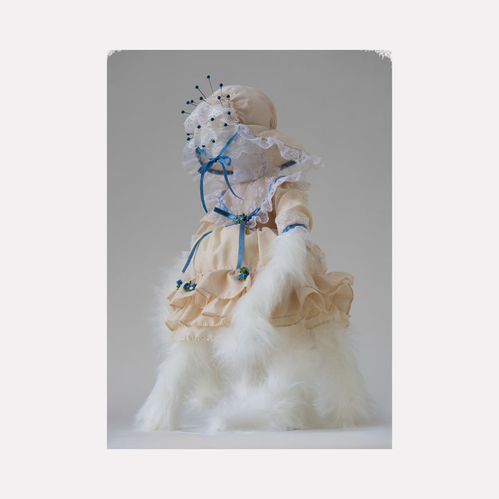 Sniffing My Own Breath by Lisyanet Rodriguez features a delicate doll-like figure dressed in soft cream fabric with intricate lace, blue ribbon accents, and floral details, wrapped in flowing white feathers, blending whimsical craftsmanship with gentle, ethereal textures in the Curina art collection.