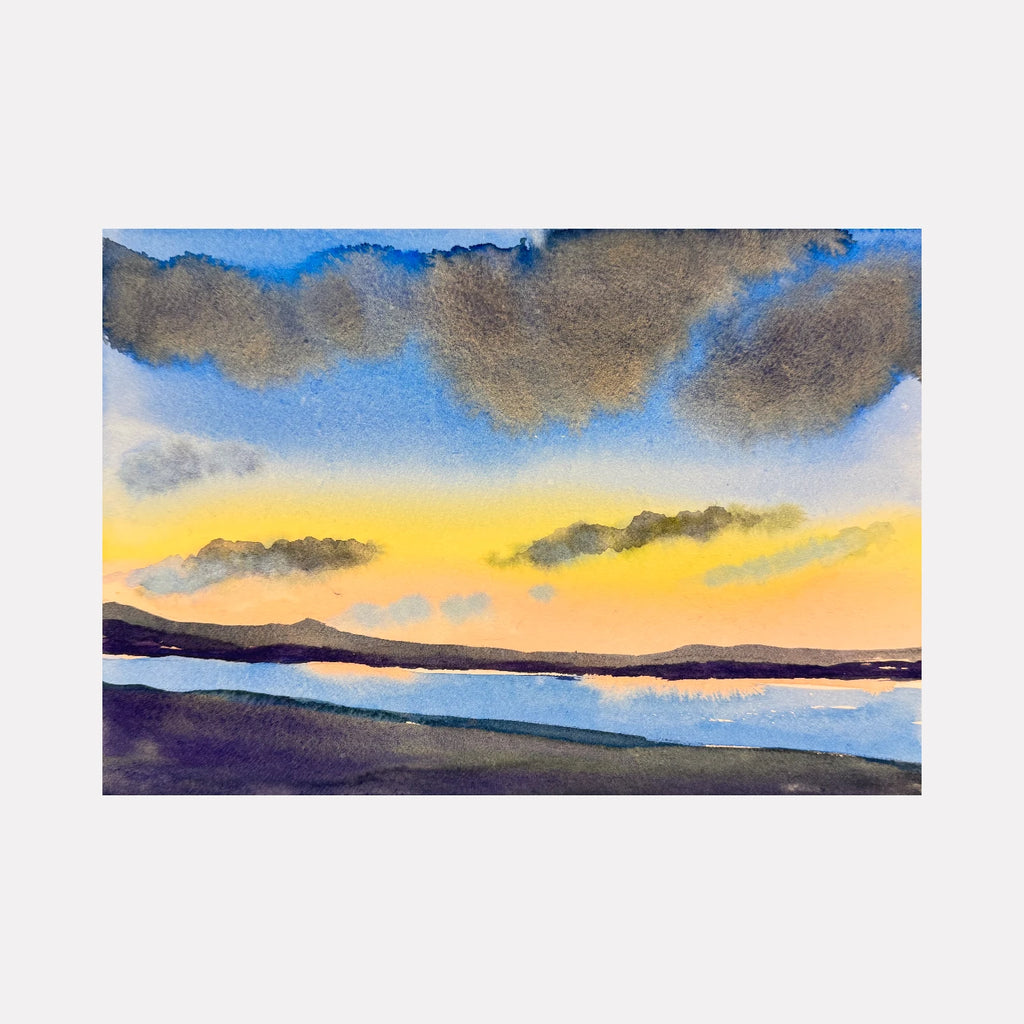 Sunset by Melanie Kozol is a Curina art piece featuring a tranquil lakeside scene with a glowing yellow and orange sky transitioning to soft blues, beneath textured clouds and distant dark hills, capturing a peaceful and reflective atmosphere in a delicate watercolor style.