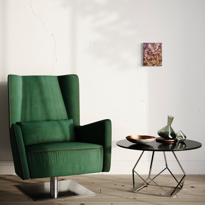 The artwork 'Deflorees Origins Gladiolus 01' displayed on a white wall features preserved flower petals in muted pink and burgundy tones arranged in an abstract collage pattern on canvas. The 8x10 inch composition, sealed with polyurethane varnish, creates a textural botanical study shown next to an emerald green armchair and modern black side table. By Valerie Hallier.