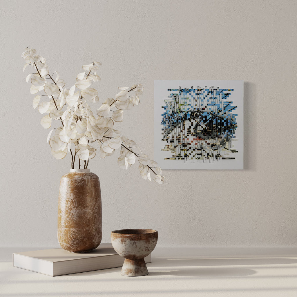 The artwork 'Sprout' by Kiana Honarmand features an intricate geometric wall piece composed of hand-cut and woven giclee prints in a 16x16 inch square format. The pattern creates a textural mosaic in shades of blue and white, with abstract pixelated elements that suggest organic movement and growth, displayed against a white wall alongside decorative white floral stems in a wooden vase. By Kiana Honarmand.
