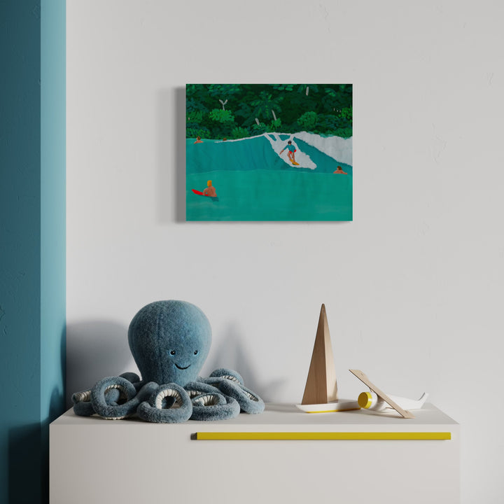 Pocket Pass by Alimo shows a surfer in red shorts riding a vibrant turquoise wave with others nearby, set against lush green tropical foliage, displayed above a white cabinet with playful decor in a minimalist room, conveying tranquil coastal art with bold, painterly style.