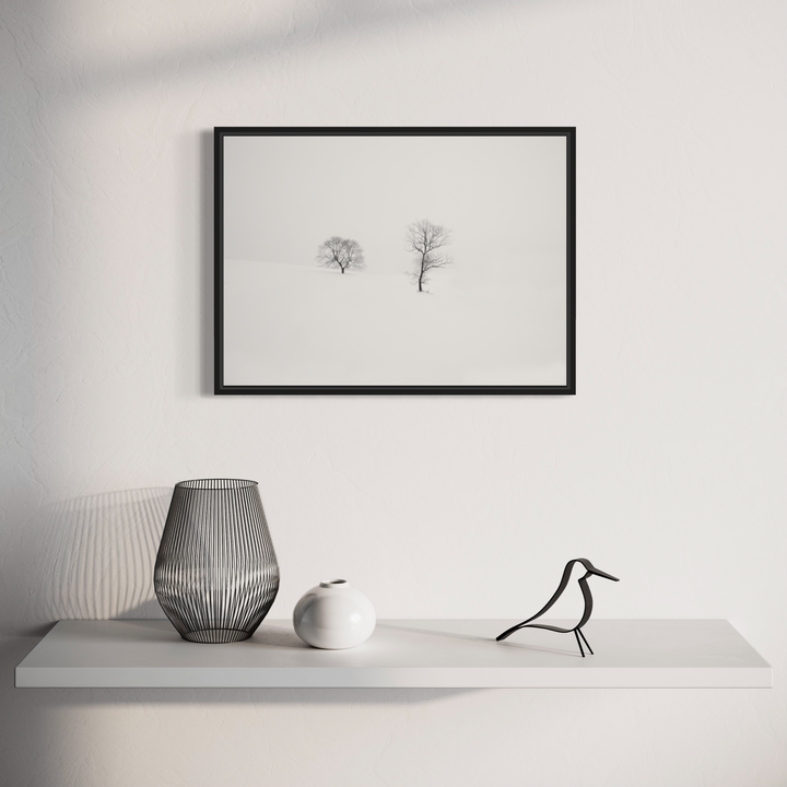 The artwork 'Rooted in Silence' by Dennis Maida features two solitary trees in a minimalist winter landscape, captured in black and white photography. The composition shows stark silhouettes against a misty white background, creating a serene and contemplative atmosphere. The trees stand as elegant forms, their bare branches reaching into the ethereal space.