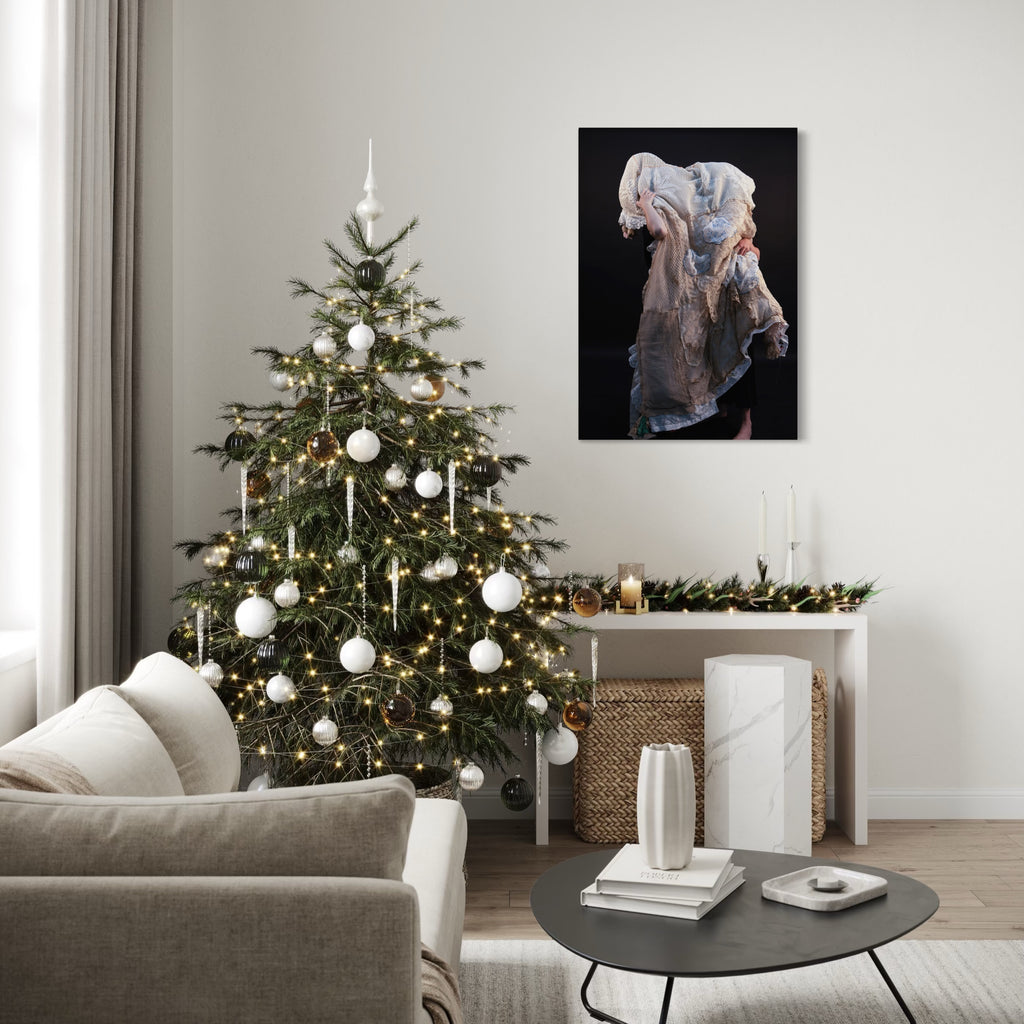 080A8374 by Eunice Pais portrays a figure cloaked in layered, textured fabric with soft, muted tones against a dark background, evoking a sense of quiet introspection. Set within a softly lit, minimalist living room adorned with festive decor, the artwork adds a contemplative and elegant presence to the serene space.
