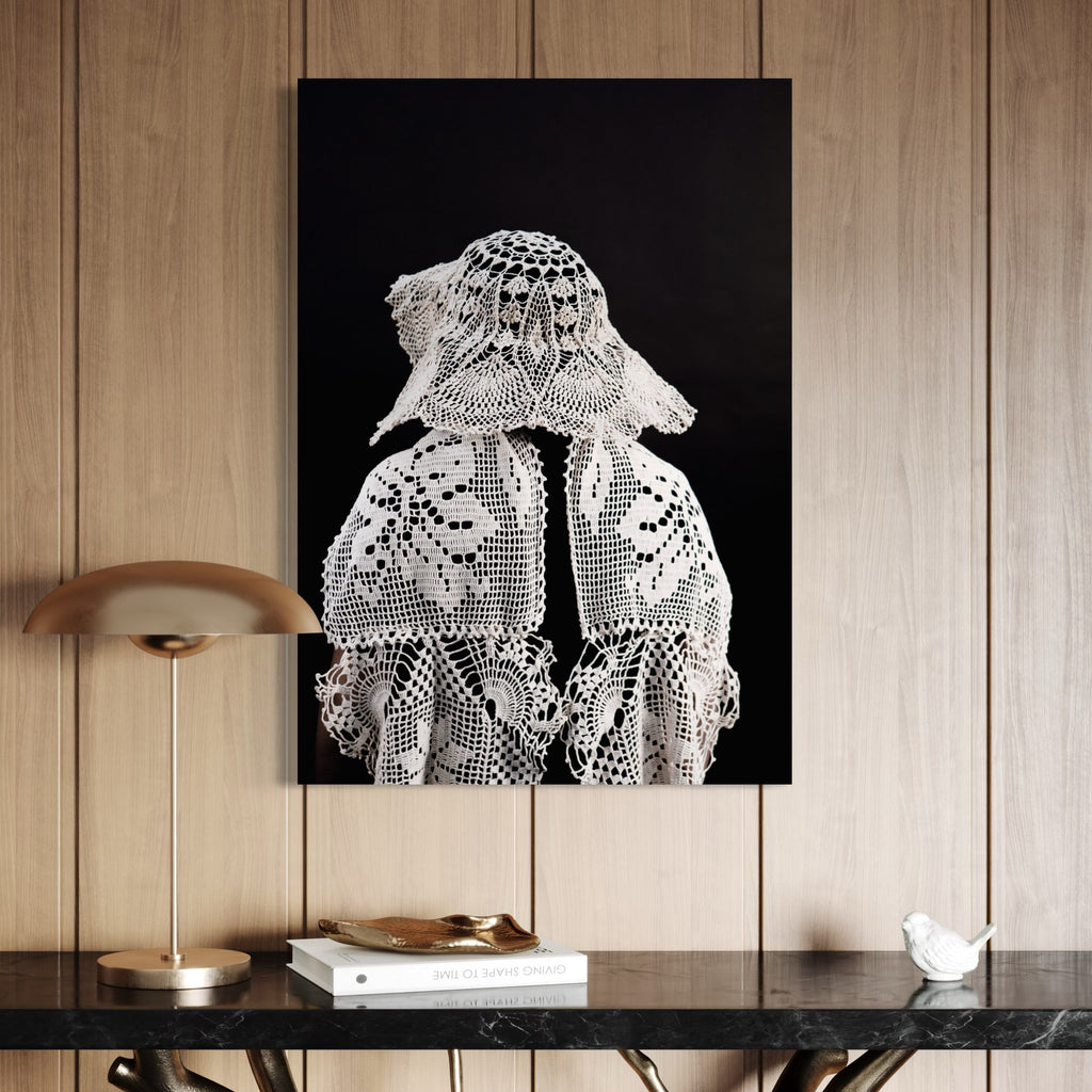 080A8515 by Eunice Pais captures a figure draped in delicate, patterned lace with a distinctive hat against a rich black backdrop. Positioned above a sleek black marble console with minimalistic decor, the artwork adds a refined, thoughtful ambiance to the warm, wood-paneled setting.