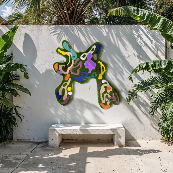 The Abstract Bouquet by Abigail Henthorne features a vibrant, organic form composed of interconnected abstract shapes in green, purple, blue, and peach tones, mounted on a white outdoor wall surrounded by tropical foliage. The piece contrasts natural surroundings with a playful, modern expression.
