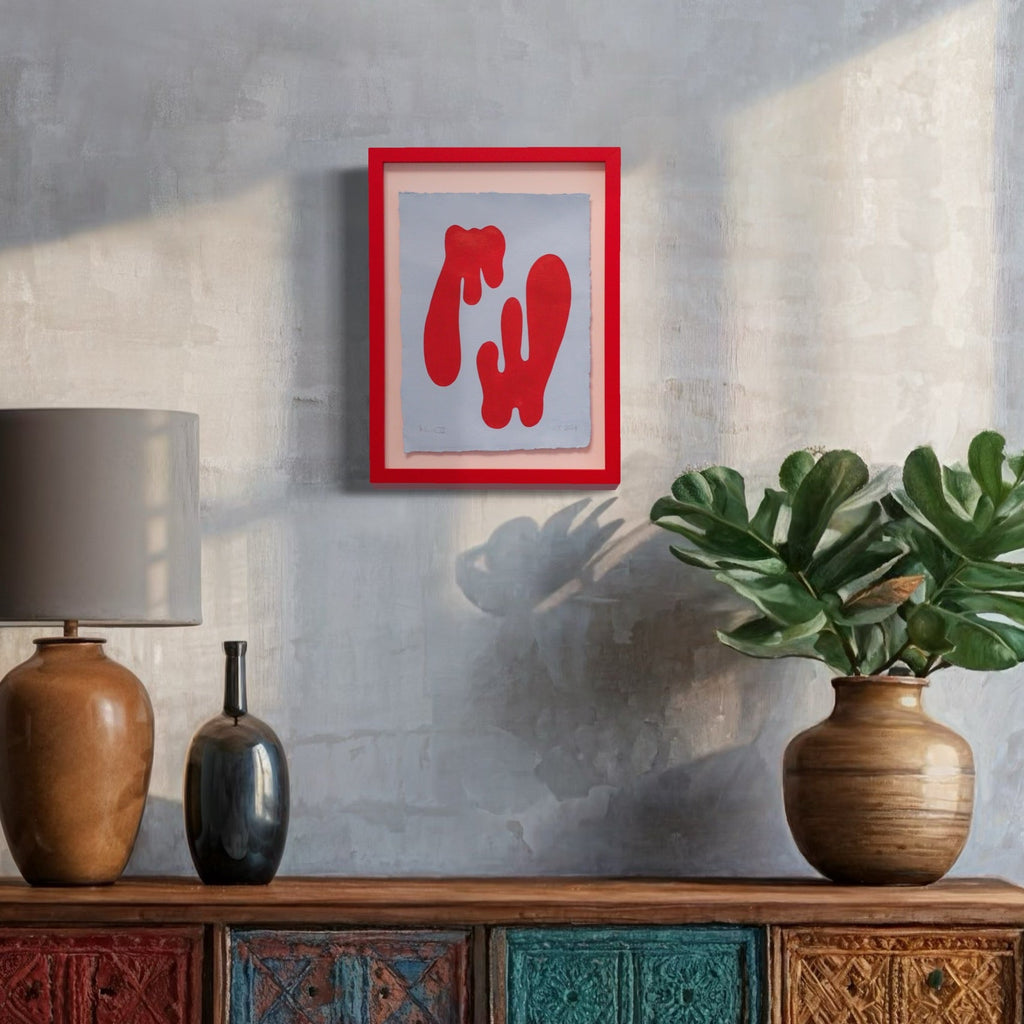 The artwork 'Balance III' by Shadia Sabagh features abstract red dripping forms against a light gray background, framed in vibrant red. Created with acrylic paint on deckle edge cotton paper, the piece displays fluid organic shapes suggesting movement and gravity. Displayed above a decorative cabinet alongside ceramic vases and monstera plants. By Shadia Sabagh.