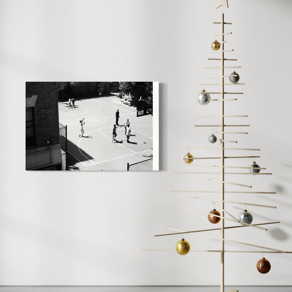 The artwork 'Ball is Life' by Cavier Coleman showcases a black and white aerial photograph of people on a basketball court, displayed on a white wall beside a minimalist wooden Christmas tree decorated with metallic ornaments. The composition captures the raw energy of street basketball in high contrast monochromatic tones, printed in 24x36 inch format.