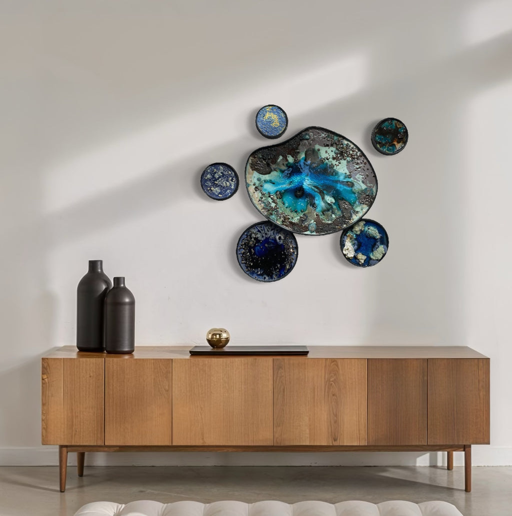 The artwork "Caribbean 2" by Lisa S. Truax features a clustered arrangement of seven circular forms in varying sizes, each displaying dynamic blue and turquoise hues with black and metallic accents. The textured surfaces evoke a fluid, cosmic quality, reminiscent of the organic abstraction in Helen Frankenthaler’s color fields and the celestial motifs found in Joan Miró’s compositions.