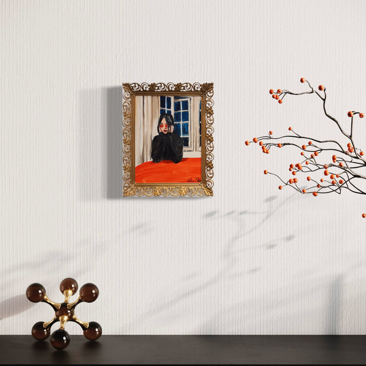The artwork 'Carina in Red Glasses' by Jillian Shea depicts a portrait in an ornate vintage metal frame with decorative gold scrollwork. A bright orange-red couch contrasts against a window backdrop. The painting features oil on canvas panel techniques with visible brushstrokes. Mounted on a white wall beside delicate cherry blossom branches and a modern molecular-style lamp. By Jillian Shea.