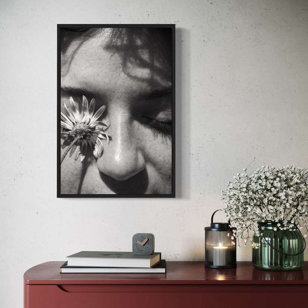 The artwork 'Communion III' by Eunice Pais features a black and white fine art photograph showing a close-up composition of a daisy flower delicately placed near closed eyes, creating an intimate and ethereal atmosphere. The monochromatic treatment emphasizes light and shadow, while the flower's petals contrast beautifully against soft facial features. The image is presented in a black frame against a white wall. Photography by Eunice Pais.